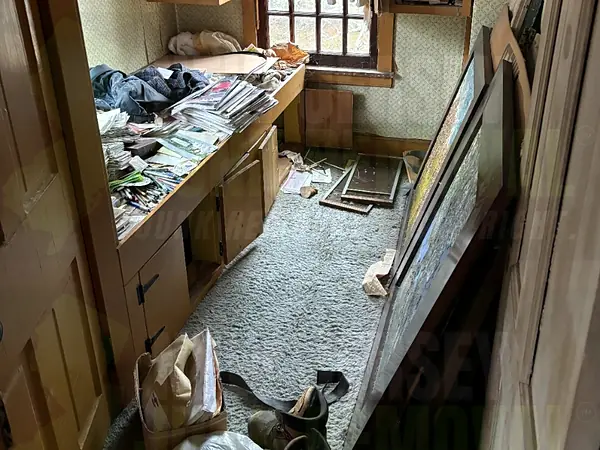 A small room in an older home, with papers, clothes and magazines piled on a built-in desk and framed pictures leaning against the wall