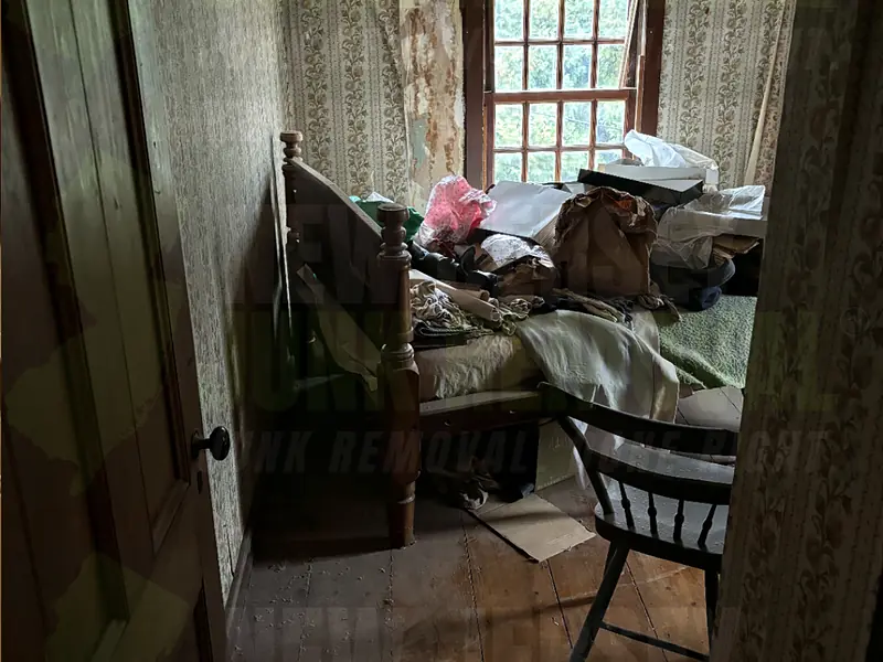 An old wooden bed piled with bedding, bags and clothes in a small bedroom before a cleanout